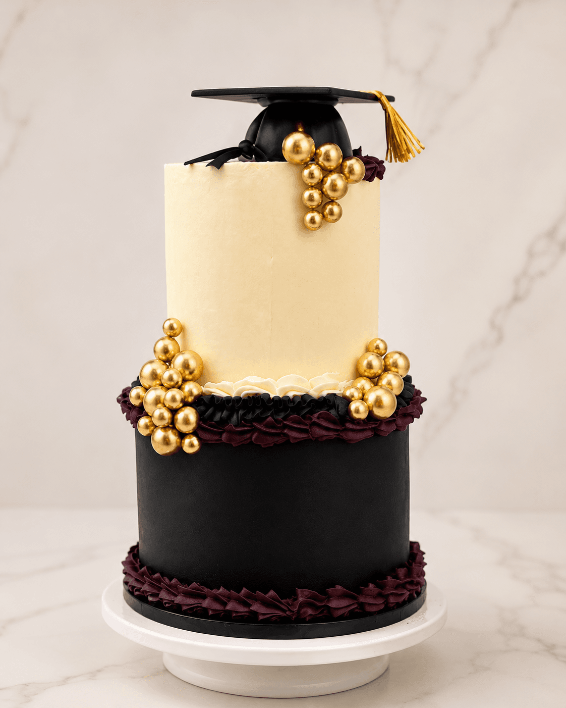 Two-tier black, white, and gold graduation cake with gold beads and a cap topper