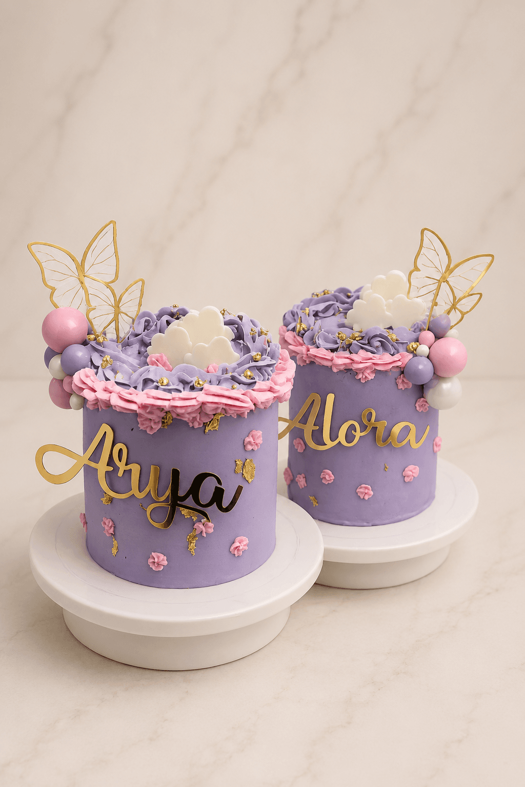 Two lilac buttercream cakes named Arya and Alora with gold butterflies, clouds, and a sphere garland