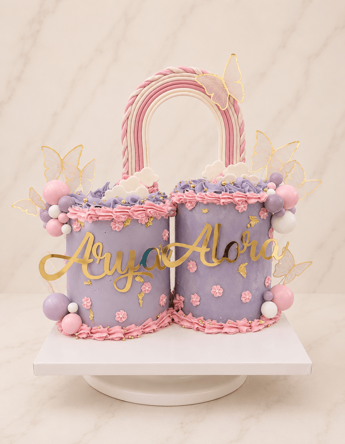 Twin lilac cakes named Arya and Alora joined by a fondant rainbow arch with gold butterflies