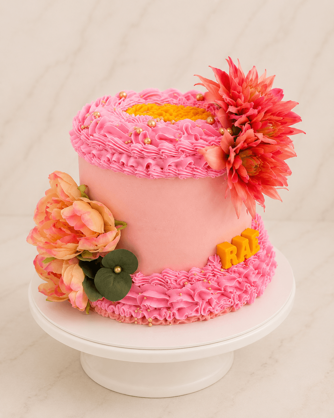 Pink ruffle buttercream cake topped with fresh dahlia and peony blooms and gold RAE letters