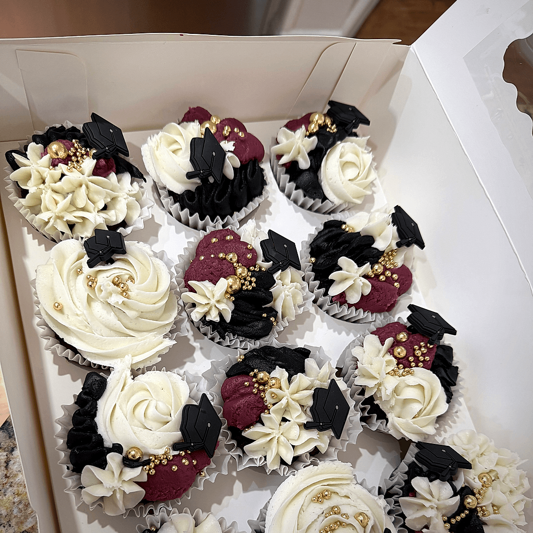 Box of graduation cupcakes in black, white, and burgundy with gold beads and cap toppers